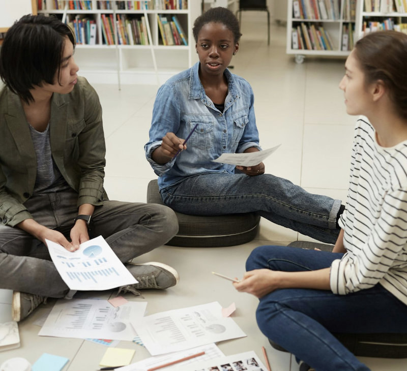 Three teachers sat on the floor in a library planning  a lesson.